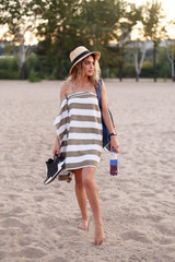 Beautiful young girl chooses a place on the beach. A girl in a towel and hat stands on the beach. Model with a briefcase, water and sneakers in his hands. Tracker on the hand. © Aleksandra