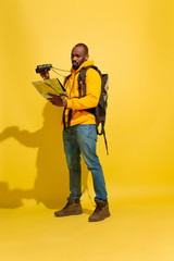 Looking forward. Portrait of a cheerful young african-american tourist guy with bag and binoculars isolated on yellow studio background. Preparing for traveling. Resort, human emotions, vacation.