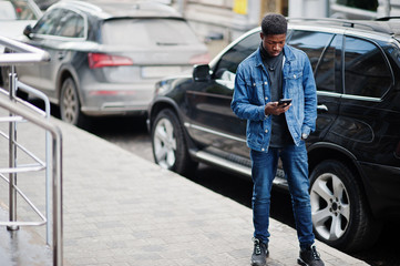 African man wear on jeans jacket posed outdoor, standing against black car with mobile phone at hand.
