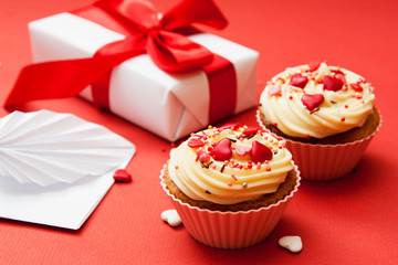 Obraz na płótnie Canvas Close-up of two cupcakes with cream and heart decor on a red background with gift and envelope.