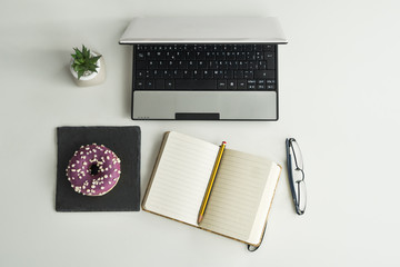 Work table with laptop, agenda, glasses, a small plant and chocolate doughnuts with shavings on a white surface. Concept of afternoon snack in the office.