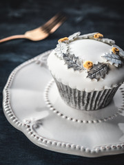 White cupcake with gold and silver decoration on white plate and dark blue background. Close up.