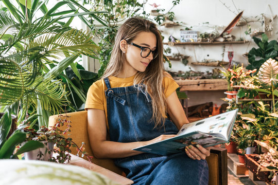 Portrait of a young woman sitting in armchair reading a book in a small gardening shop - Powered by Adobe