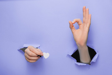 Close up of female hands holding little white hearts through and shows an ok sign a torn purple...