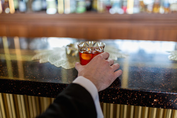 Rich and successful businessman holds a glass of Scotch whisky with an ice cube on a wooden table, sitting at the bar . Hand holding liquor whiskey or cognac in the glass on the background of the bar