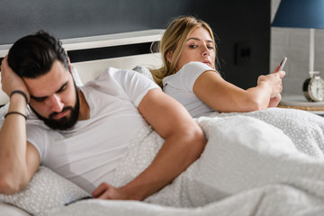Young adult couple using smartphones in the bed addicted to social networking. Mobile addiction concept. Jealous wife.