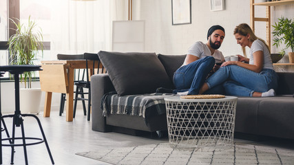 Young hipster couple sitting on sofa at home talking and drinking coffee