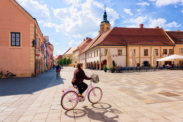 Fototapeta premium Senior woman on bicycle at Street cafes and restaurants on Town Hall on King Tomislav Square in Old city of Varazdin in Croatia. Cityscape of famous Croatian town in Europe in summer