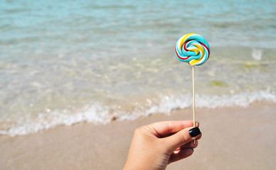 Female hand holds colorful candy on sea background.