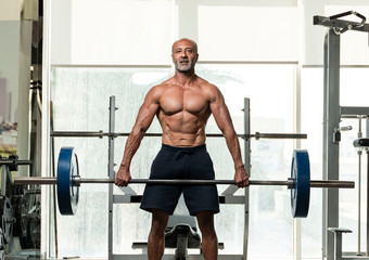 Fototapeta na wymiar A strong muscular shirtless mature older bodybuilding athlete with balding gray hair holding a heavy barbell, looking at camera in a gym