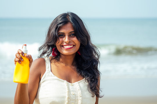 Portrait Of A Beautiful And Smiling Snow-white Smile Indian Woman Black Curly Hair And Dark Skin In A White T-shirt Holding Bottle Of Sunscreen Spray On Beach.girl Enjoying Spf Body Paradise Vacation