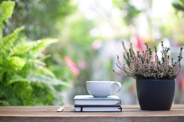 White coffee cup and notebooks with heathers plant in black pot on wooden table at outdoor