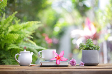 White coffee cup and pink flower with plant and white teapot on wooden table at outdoor