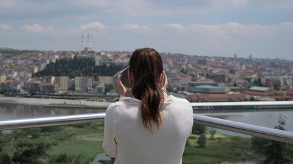 Fototapeta na wymiar brunette woman in white sweater looks through binoculars exploring cityscape stretching along bay backside view
