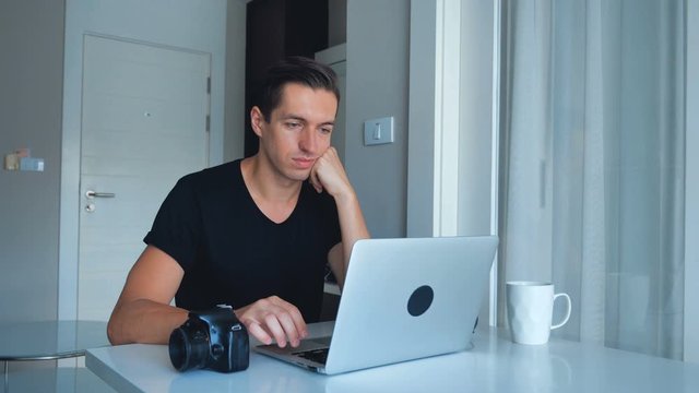 Young photographer using laptop processing images on his laptop and drinking coffee at home. Camera, External Hard Drive Lie Beside.