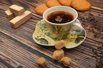 A Cup of black tea, tea leaves, pieces of brown sugar, oatmeal cookies, waffles on a wooden background. Close up.