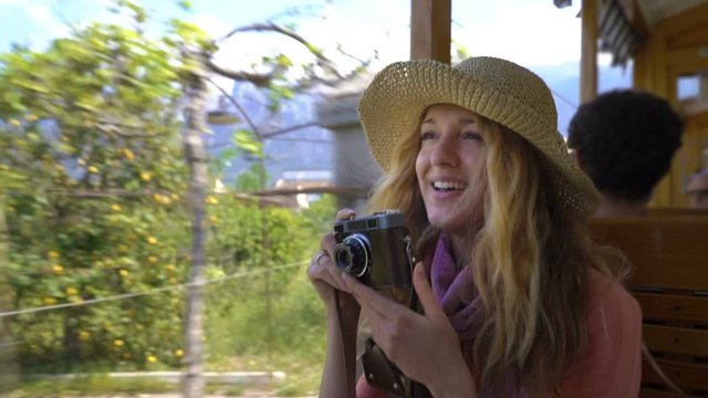 A young woman wearing straw hat enjoying traveling on an old tram or train, taking pictures of beautiful tourist locations using vintage camera, feeling excited and happy