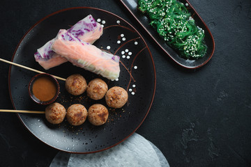 Nem nuong or fried vietnamese meatball skewers with spring rolls and seaweed salad, top view over black stone background
