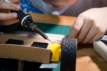 Closeup, hands of tween / teen solder his simple car robotics for STEM project, right hand hold electric soldering iron, left hand hold lead-free fusible metal alloy.