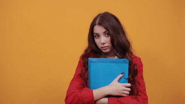 Serious brunette young woman in fashion white shirt and red jacket keeping blue folder, looking at camera isolated on orange background in studio. People sincere emotions, lifestyle concept.