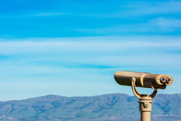 Coin operated binocular telescope for distant viewing. Blurred range of green hills, mountains in the distance under cloud sky
