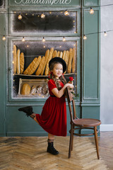 A happy smiling six-year-old girl in a red velvet dress and black beret outside a baguette bakery holds a red rose in her hand