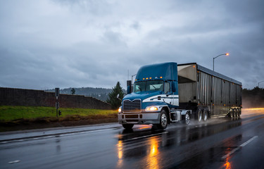 Big rig bonnet blue semi truck transporting cargo in covered bulk semi trailer running on the wet glossy road with raining weather evening