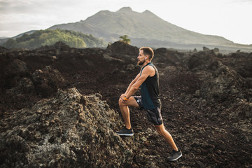 Young hipster runner with beard stretching and warming-up for trail running outdoors. Listening...