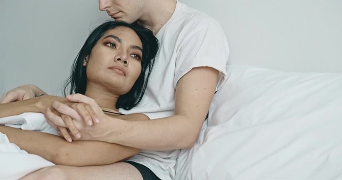 Lover sitting in bed thinking about future, tilt shot. Caucasian male with his girlfriend, asian woman in black lingerie. Happy lover in bed concept.