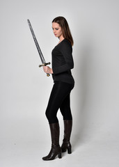 Fototapeta na wymiar full length portrait of a pretty brunette girl wearing a black shirt and leather boots, holding a sword. Standing pose facing away and holding a sword, on a grey studio background.
