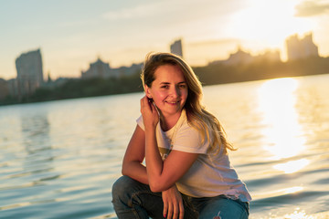 Young brunette smiling woman posing at the river shore at the sunset with cityscape at the background