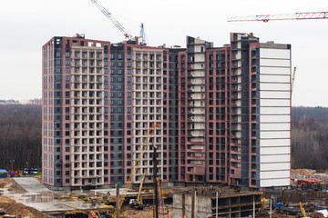 Under construction high-rise house near the forest with yellow construction cranes and with builders