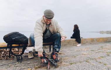 An elderly caucasian man in roller skates, a down jacket, a cap and glasses sits on a bench and...