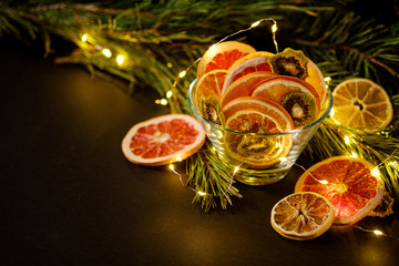 Fototapeta na wymiar Creative moody holiday Christmas New Year food fruits with dried grapefruit, kiwi, orange and lemon in glass bowl with branch of fir tree with warm led lights, angle view, copy space