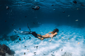 Free diver woman with fins glides over sandy bottom with fishes in transparent sea