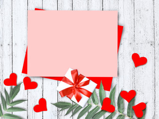 Mock up greeting composition with pink empty blank, red hearts, gift and palm leaves on wooden background. Valentines day, Mothers day or wedding flatlay with copyspace.