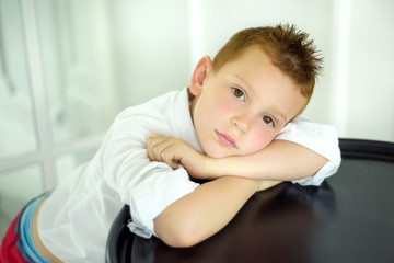 Portrait of 7 years old caucasian boy in white shirt with his hands near face. Fashionable good looking little boy looking in camera. Stylish trendy schoolboy in light room.