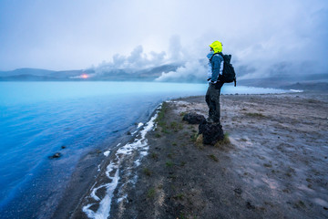 Man with hiking gear stands on shore looking towards Geothermal powerplant at the blue lagoon in Jarabodin/Myvatn in Iceland during blue hour.