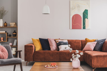 Real photo of a cozy, brown corner sofa with decorative cushions standing in a bright living room interior with an abstract painting - 313242501