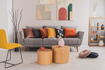 Real photo of round, wooden tables standing in front of a gray couch with colorful pillows in bright living room interior