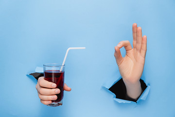 Close up of female hand holding a glass of fresh red juice with a straw through torn blue paper wall. Fruit drinks and healthy nutrition. Hand sign. Hole in wall. Like gesture