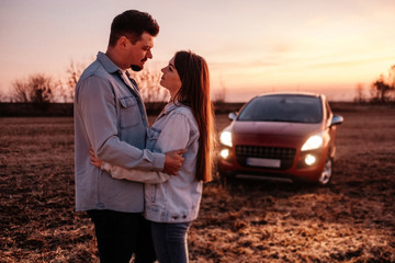 Young Happy Couple Dressed Alike in White Shirt and Jeans Enjoying Road Trip at Their New Car, Beautiful Sunset on the Field, Vacation and Travel Concept