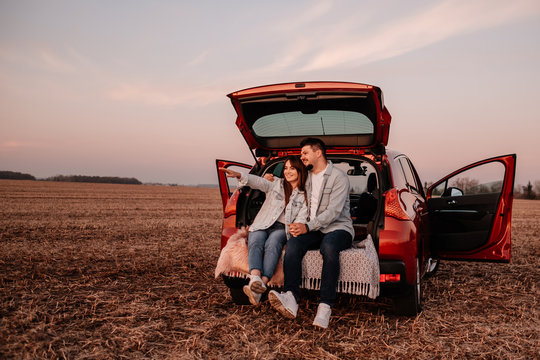 Young Happy Couple Dressed Alike in White Shirt and Jeans Sitting at Their New Car Trunk, Beautiful Sunset on the Field, Vacation and Travel Concept