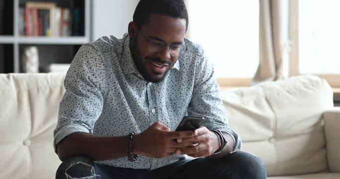 Smiling african guy using smart phone sitting on sofa