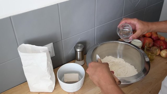 Mediterranean Man Hand pour a glass of water over white flour inside metal bowl. Mixing with wooden spoon for Homemade Pizza dought recipe, modern minimalist background.