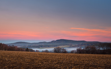 Fototapeta na wymiar landscape at sunset, in the foreground field and trees, in the valley white fog, in the background forest and mountains, red and blue sky