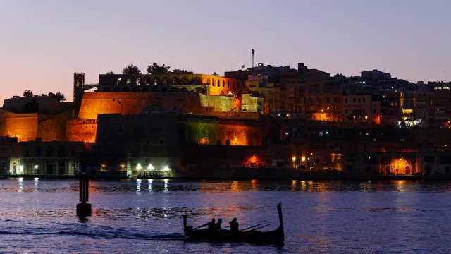 City of Valletta at dusk in Malta, small traditional luzzu boat with tourists on sightseeing tour in the Grand Harbour.
