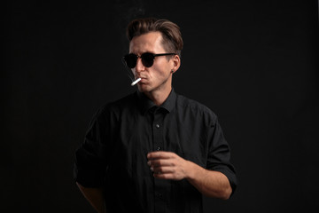 Stylish young man in a black t-shirt and sun glasses smoking a cigarette over black background.