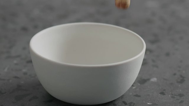 Slow motion roasted hazelnuts falling into white bowl on terrazzo countertop