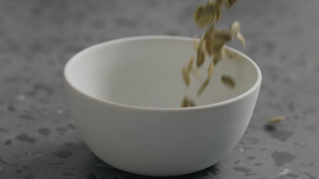Slow motion dried pumpkin seeds falling into white bowl on terrazzo countertop
